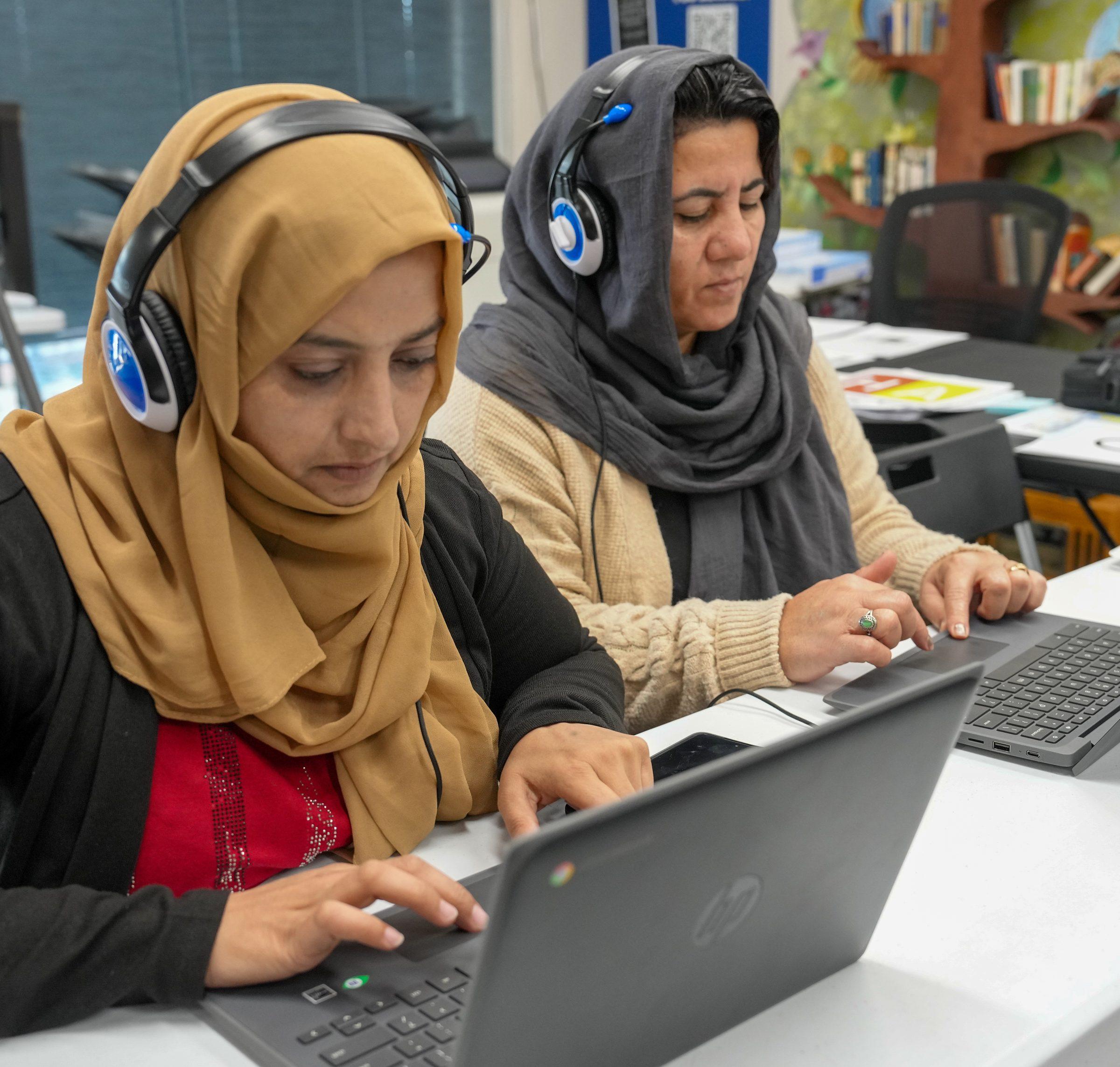 Two community members wearing headphones and working on laptops together in a classroom.